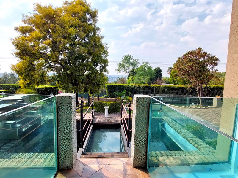 A well-maintained outdoor pool area featuring a modern glass balustrade, mosaic-tiled pillars, and a central bridge over a sunken water feature.