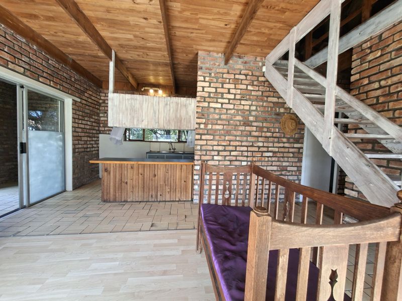 Open plan living area featuring exposed brick walls, timber ceiling, a wooden staircase, and a functional kitchen area with built-in cabinetry.