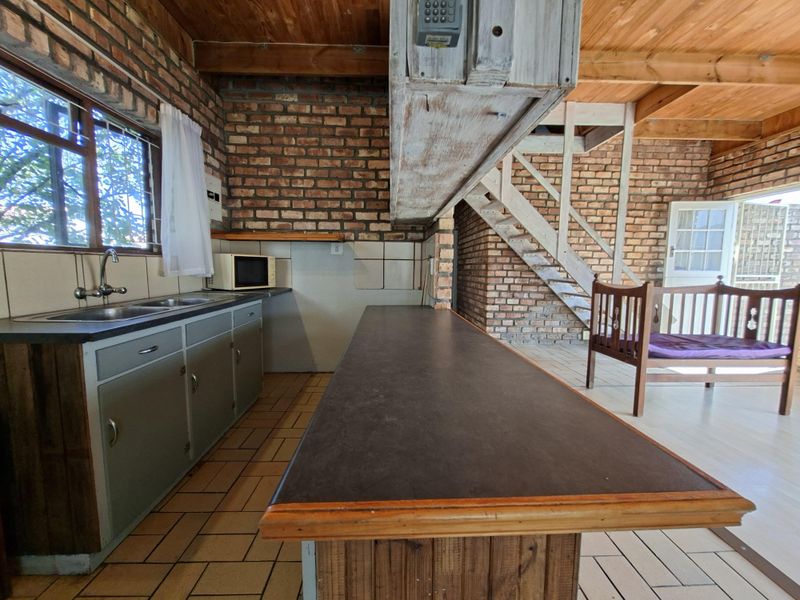 Open plan kitchen and living area featuring exposed brick walls, timber ceiling, tiled flooring, and a wooden staircase leading to an upper level.