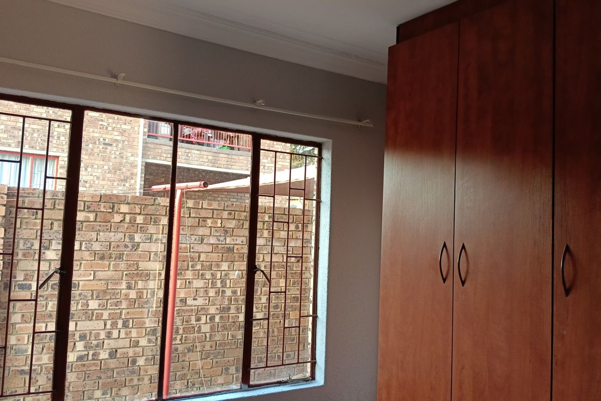A bedroom featuring a built-in wooden wardrobe and large windows fitted with security bars, offering a view of the exterior brick wall.