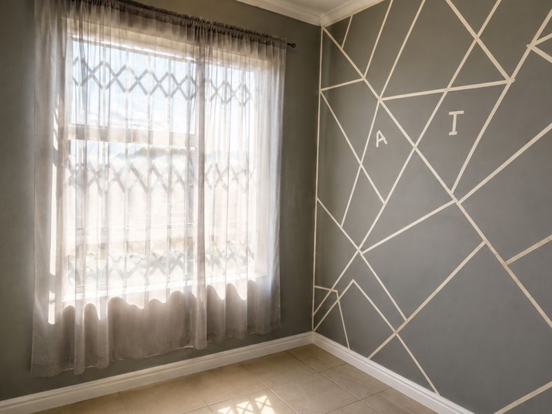 A bright, empty bedroom featuring tiled flooring, a window with a fitted security gate, and a decorative geometric accent wall.