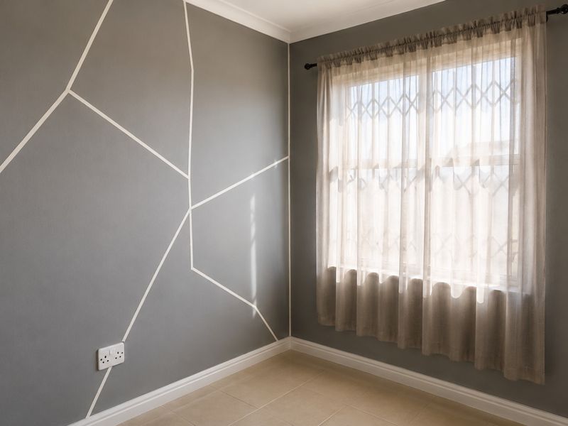 A clean, empty bedroom featuring tiled flooring, a security gate on the window, and a modern geometric accent wall.