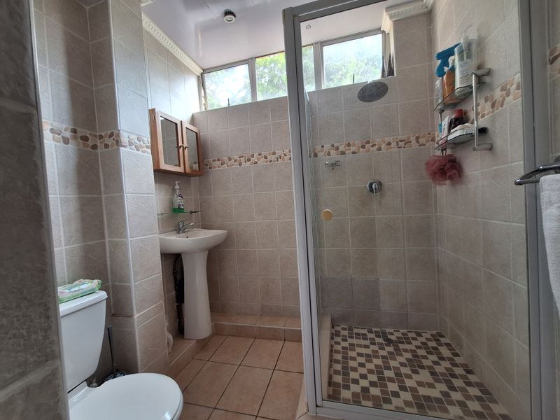 A functional bathroom featuring a pedestal basin, a glass-enclosed shower, and neutral tiled finishes with high-level windows for natural light.
