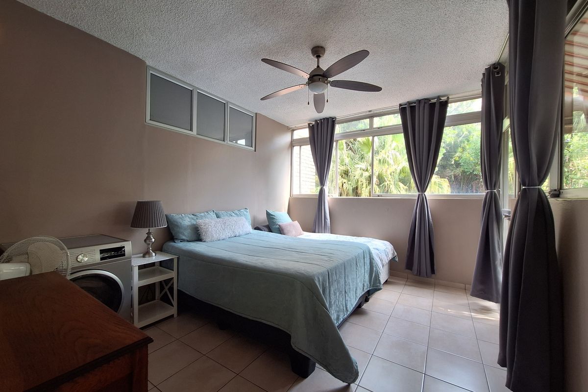 A bedroom featuring tiled flooring, a ceiling fan, and large windows providing natural light and garden views.