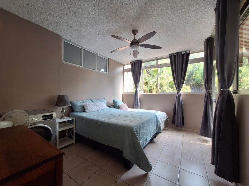 A bedroom featuring tiled flooring, a ceiling fan, and large windows providing natural light and garden views.