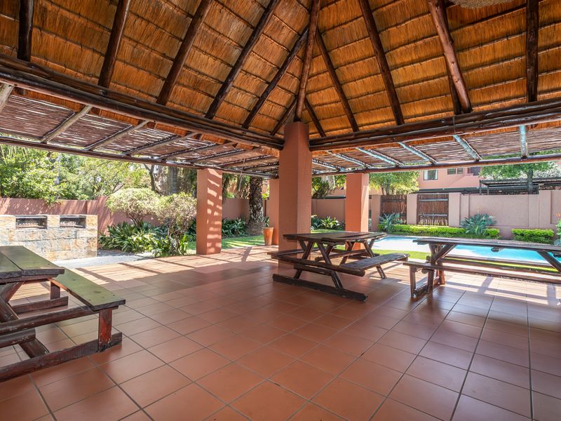 Covered outdoor patio area featuring a thatched roof, tiled flooring, and a built-in braai, overlooking the swimming pool and garden.