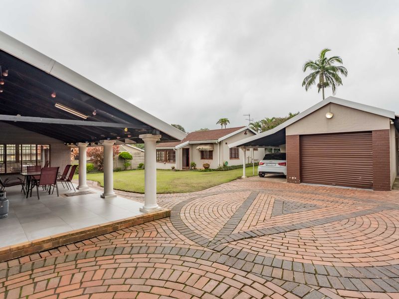 Spacious rear exterior featuring a covered patio with tiled flooring, a paved driveway, a garage, and a carport set within a landscaped garden area.