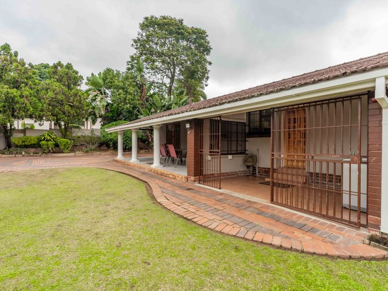 Rear view of the house featuring a covered patio area, brick paving, and a well-maintained lawn with established garden surroundings.