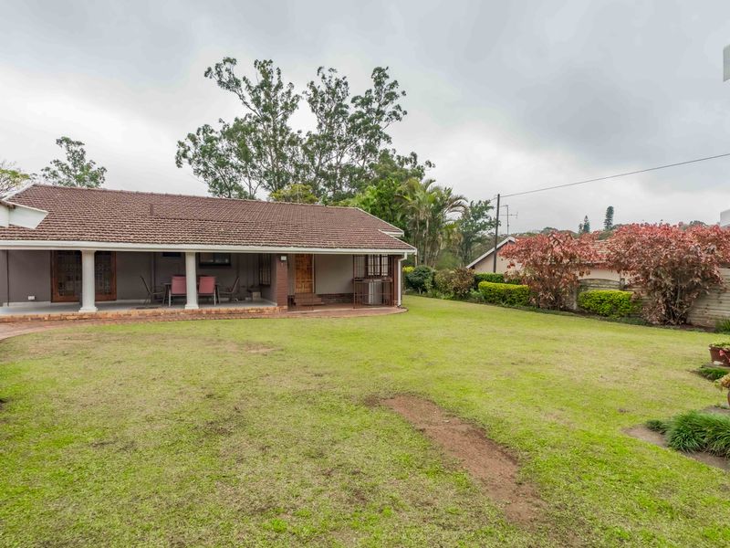 A view of the rear exterior featuring a covered patio, brick paved area, and a spacious lawn with a boundary wall.