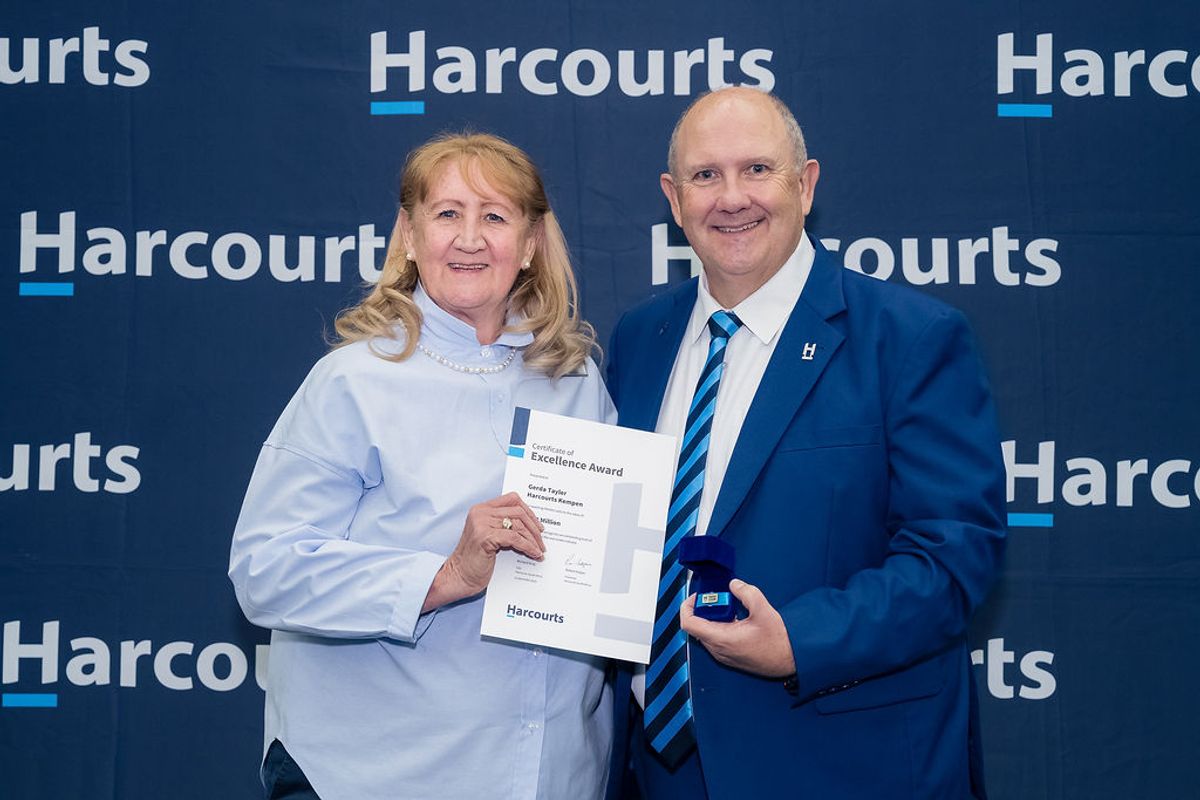 A photograph of two people holding an award certificate in front of a branded backdrop.