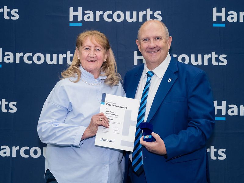 A photograph of two people holding an award certificate in front of a branded backdrop.