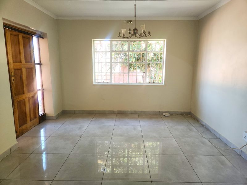 A bright, empty living room featuring neutral tiled flooring, a wooden entrance door, and a window fitted with security bars.