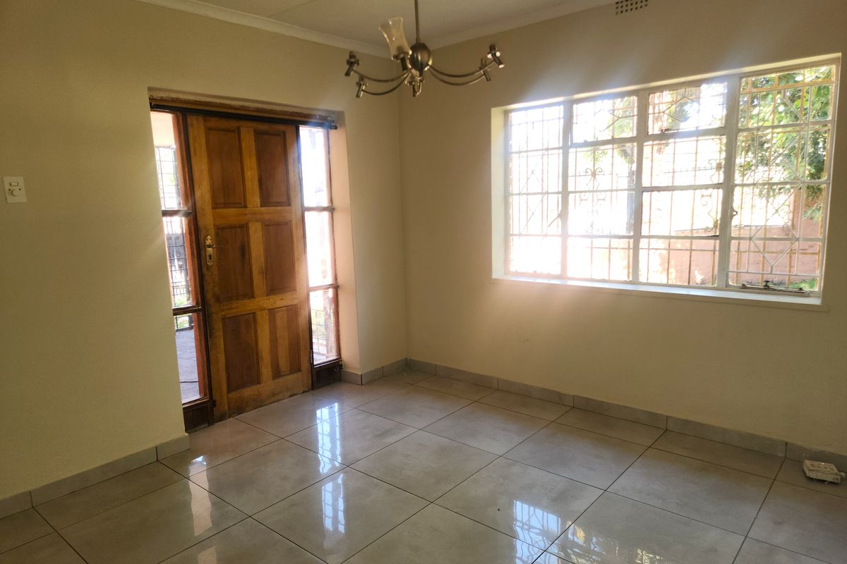 A bright, empty living area featuring tiled flooring, a wooden entrance door, and large windows with security bars.