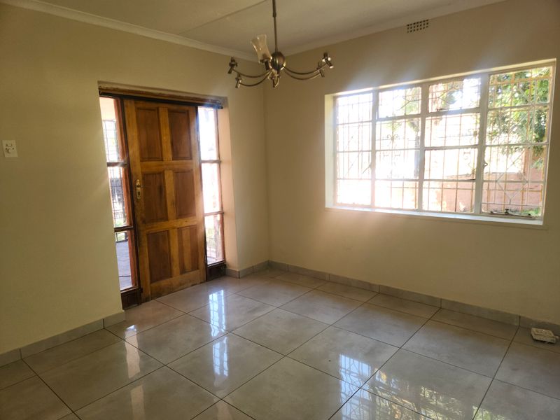 A bright, empty living area featuring tiled flooring, a wooden entrance door, and large windows with security bars.