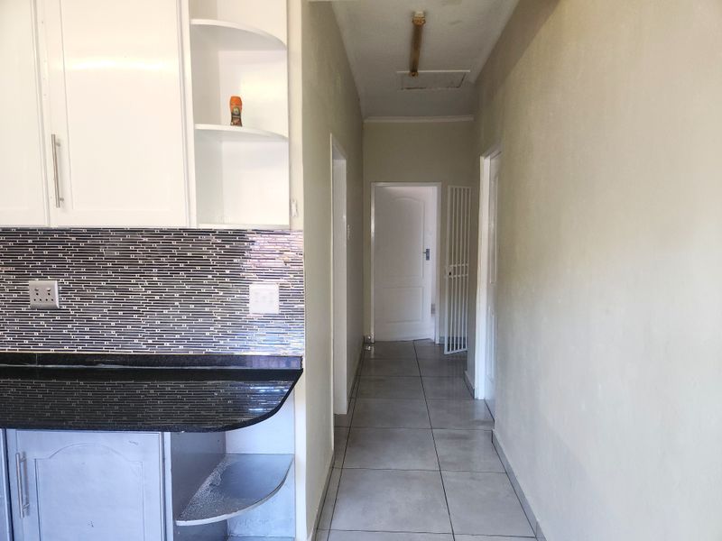 A functional kitchen area featuring built-in cabinetry, granite countertops, and a mosaic tile backsplash, leading into a tiled hallway.