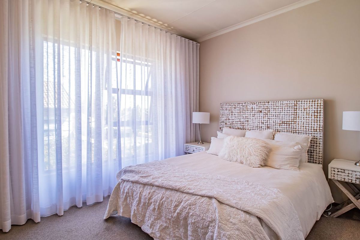 A bright and well-maintained bedroom featuring neutral wall tones, carpeted flooring, and a large window with floor-to-ceiling curtains.