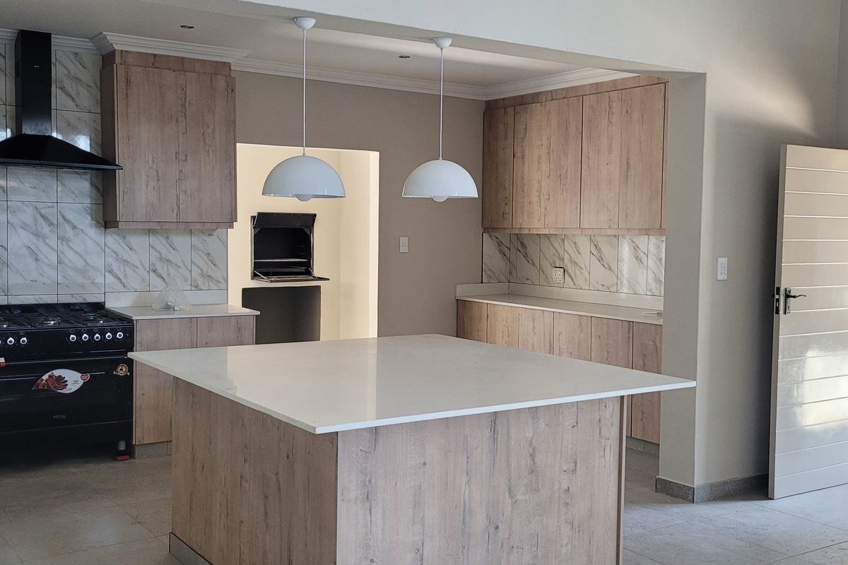 Modern kitchen featuring wood-finish cabinetry, stone countertops, a central island, gas stove, and a built-in braai area in the adjacent scullery space.