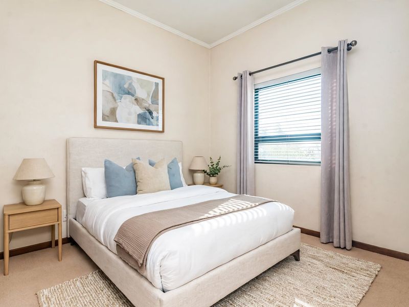 A well-maintained bedroom featuring neutral wall tones, carpeted flooring, and a window with fitted blinds and curtains.