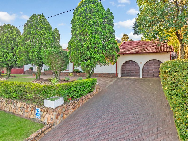 Front view of the property featuring a double garage, brick paved driveway, and established garden with stone retaining walls.