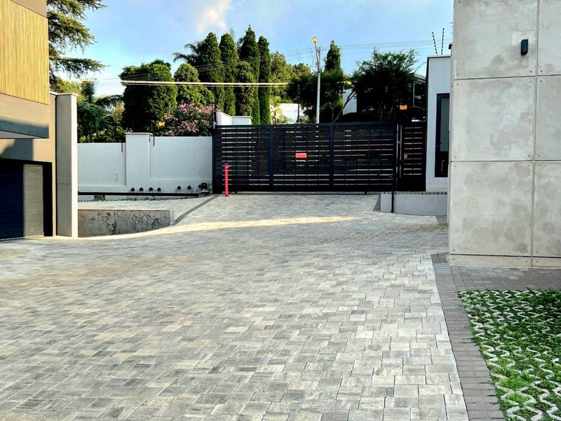 A modern paved driveway leading to a secure metal gate, featuring contemporary concrete architectural elements and integrated grass pavers.
