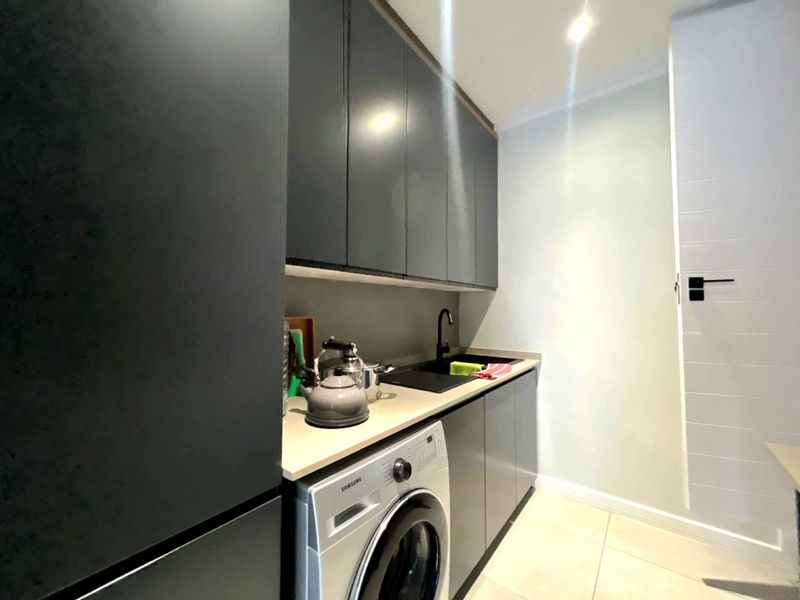 Modern laundry room featuring sleek dark cabinetry, a stone-look countertop, integrated sink with black fixtures, and tiled flooring.