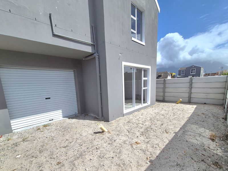 Rear exterior view of the property featuring a roller garage door, sliding glass door access, and an unfinished sandy backyard with boundary walling.