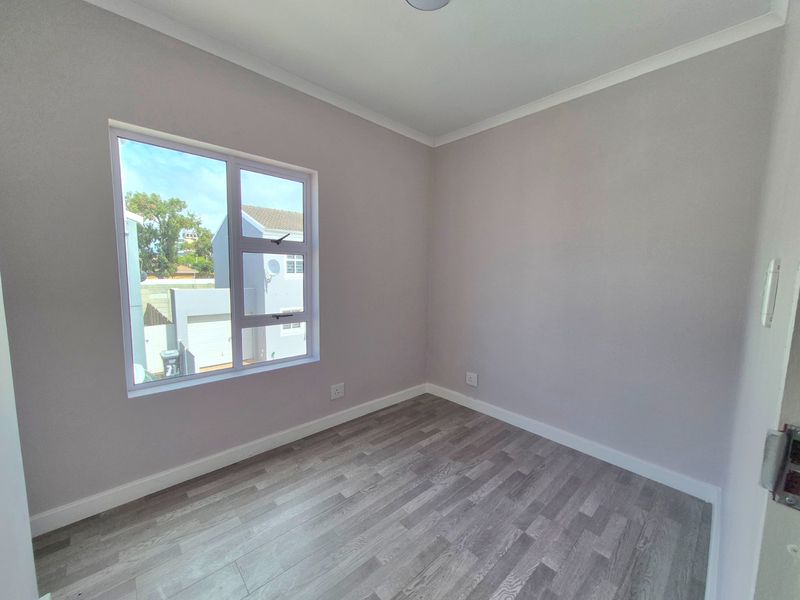 A clean, empty bedroom featuring modern grey laminate flooring, neutral wall paint, and an aluminium-framed window providing natural light.