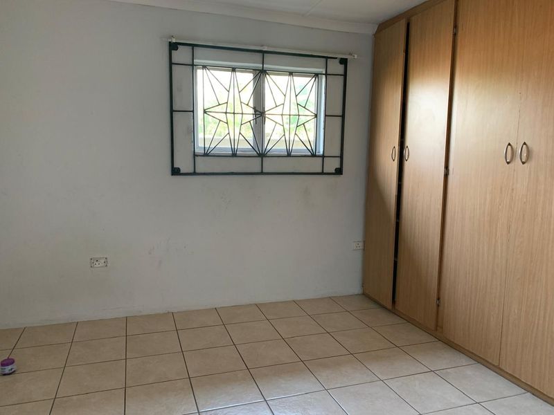 A functional bedroom featuring tiled flooring, built-in wooden cupboards, and a window fitted with security bars.