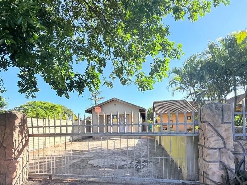 Front view of a cottage featuring a gated entrance, concrete driveway, and stone-clad pillars, set within a residential property with established trees.