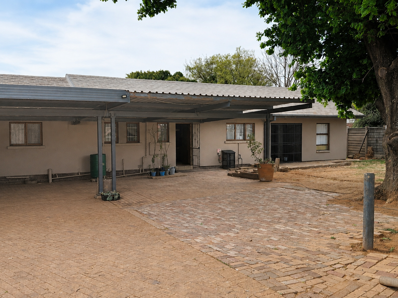 Rear exterior view featuring a large paved driveway, extensive carport structure, and mature tree in a spacious backyard setting.