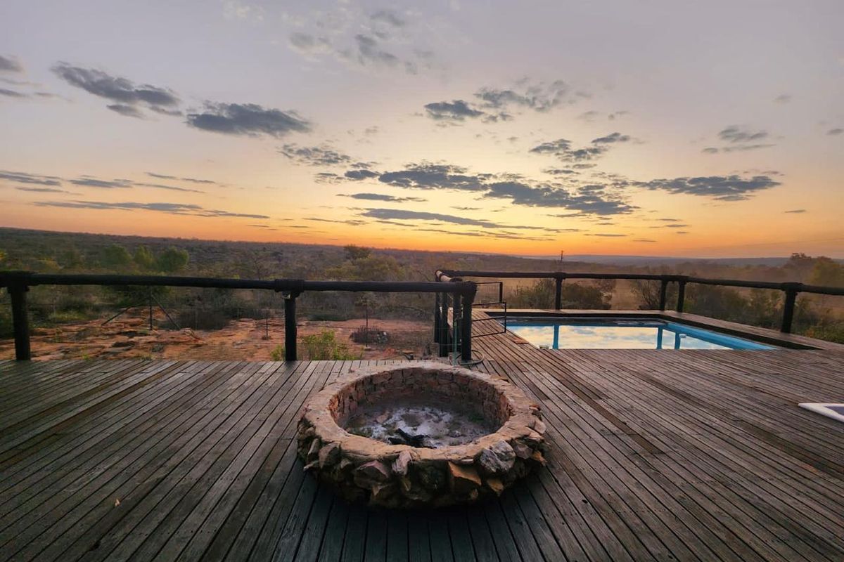 Outdoor timber deck featuring a built-in stone fire pit and swimming pool, overlooking a vast natural landscape at sunset.