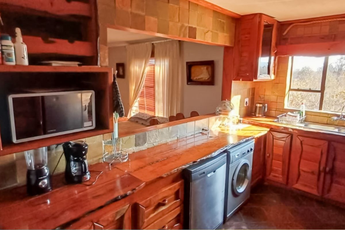 A rustic kitchen featuring wooden cabinetry, stone tile backsplash, and wooden countertops, with a view through to the adjacent living area.