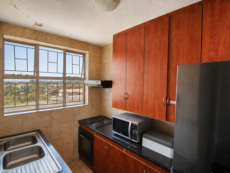 Compact kitchen featuring wood-finish cabinetry, dark countertops, built-in oven and hob, and tiled surfaces. Ceiling requires repair due to visible plaster damage.