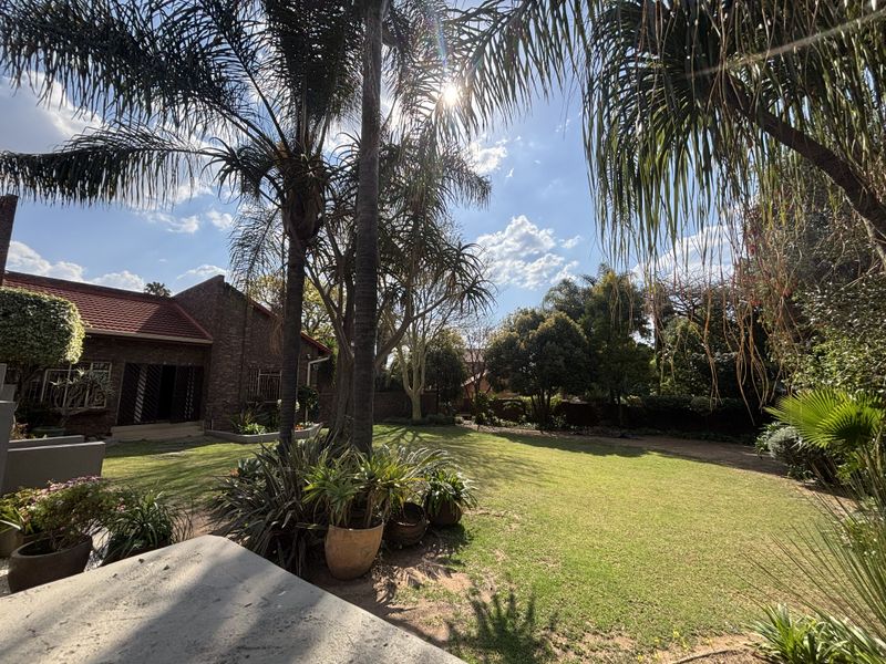 A spacious, well-maintained garden featuring mature trees, lush lawn, and a view of the brick-built house exterior.