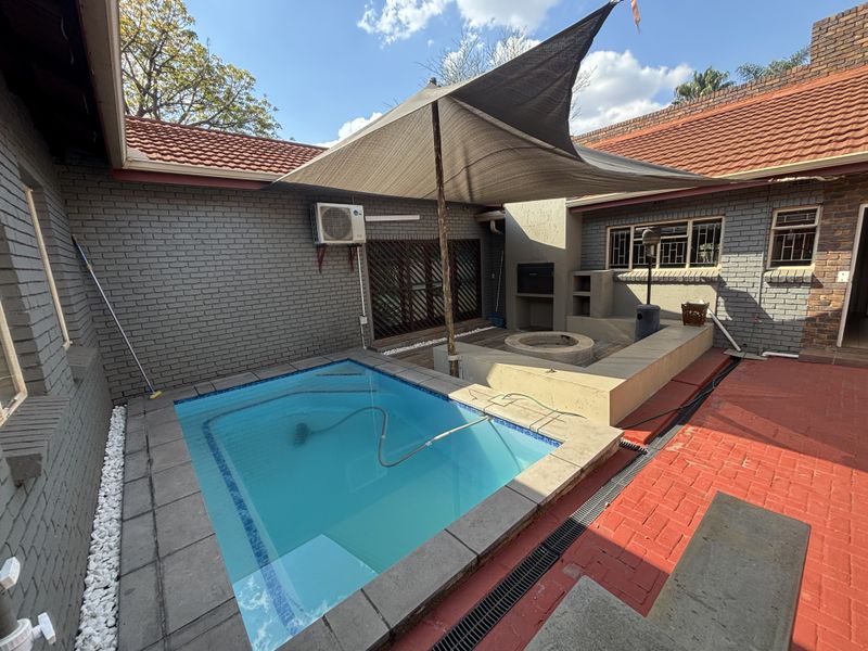 Outdoor entertainment area featuring a swimming pool, built-in braai, fire pit, and shade sail, set within a private courtyard with brick paving.