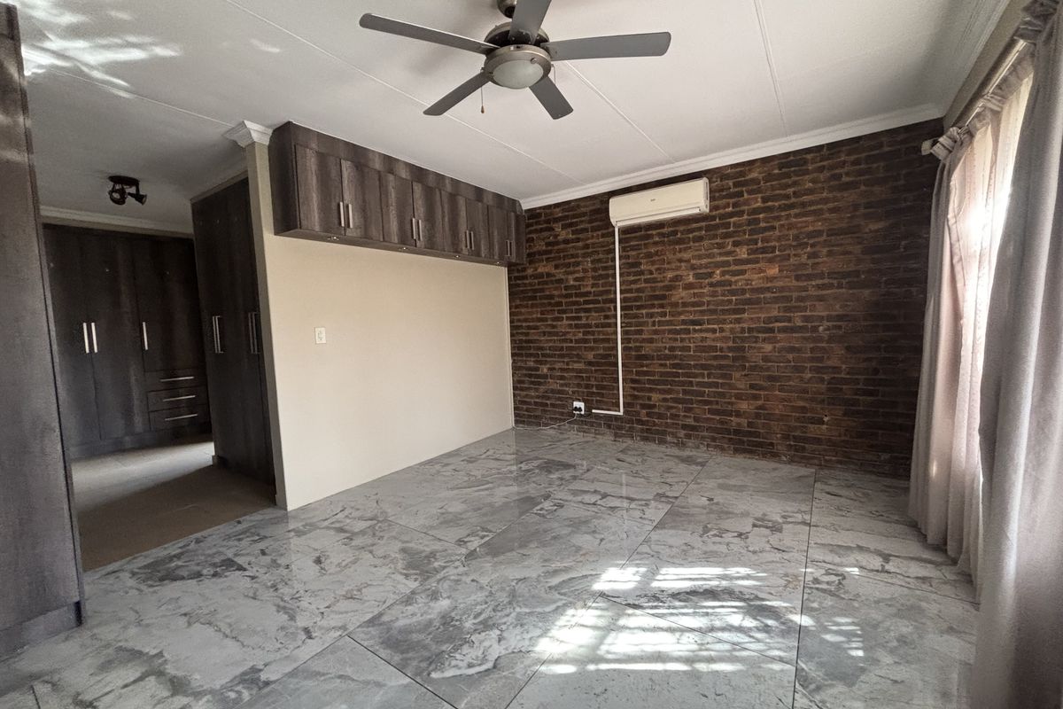 Spacious bedroom featuring polished marble-look floor tiles, an exposed brick feature wall, built-in cabinetry, ceiling fan, and a wall-mounted air conditioning unit.