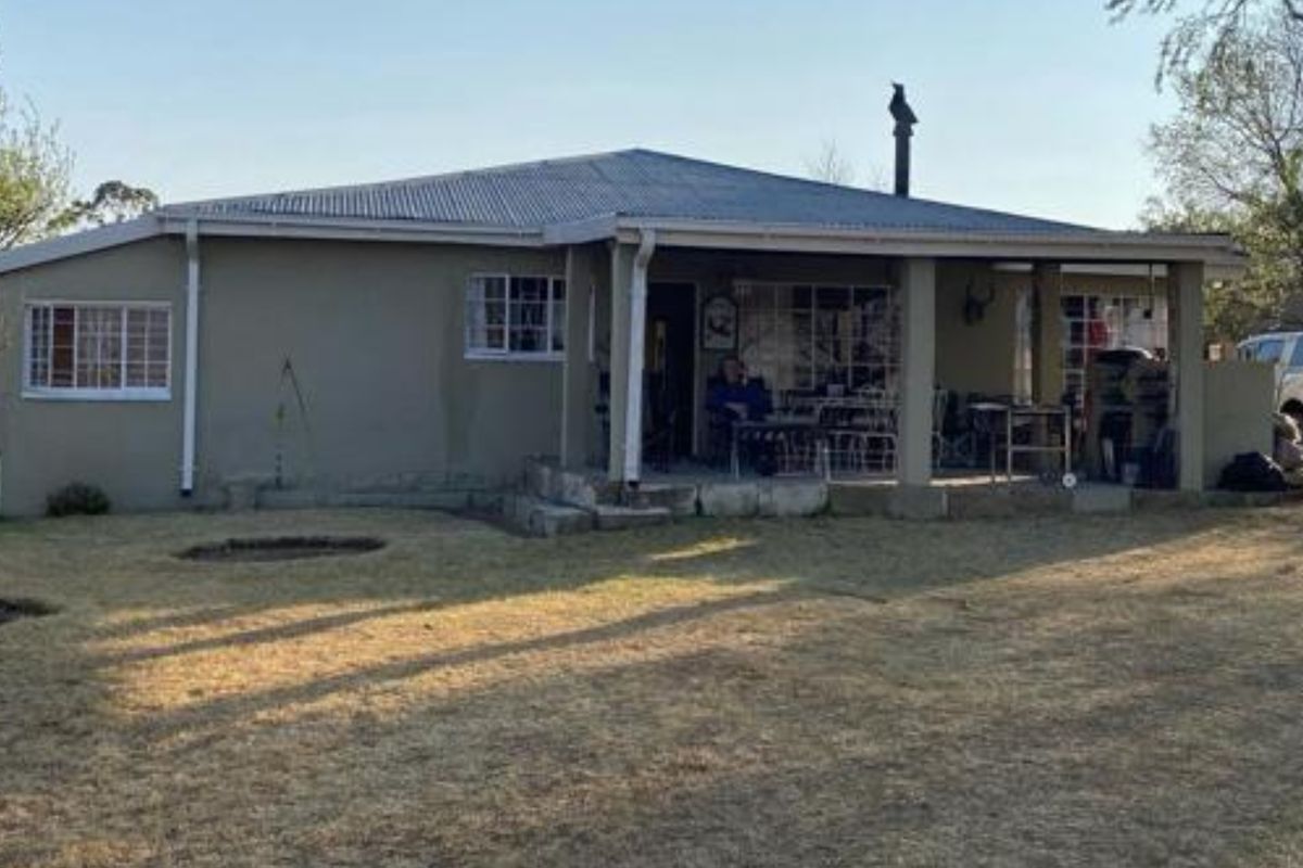 Rear view of a single-story house featuring a covered patio area, corrugated metal roofing, and a spacious surrounding garden.