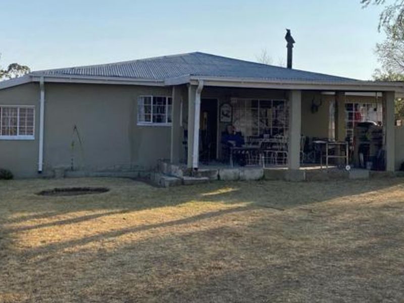 Rear view of a single-story house featuring a covered patio area, corrugated metal roofing, and a spacious surrounding garden.
