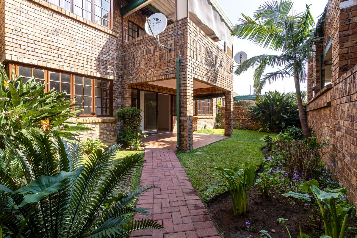 A private rear garden featuring a paved walkway, established landscaping, and a covered patio area attached to the face brick townhouse structure.