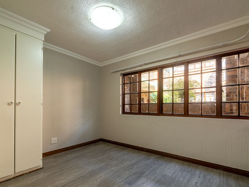A bright, empty bedroom featuring laminate flooring, a built-in wardrobe, and large timber-framed windows with a view of the exterior brickwork.