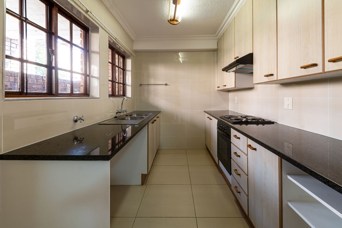 A clean, functional galley-style kitchen featuring granite countertops, built-in oven, gas hob, extractor fan, and ample wooden cabinetry with tiled flooring.