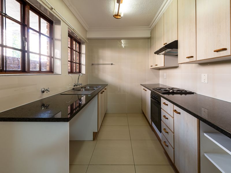 A clean, functional galley-style kitchen featuring granite countertops, built-in oven, gas hob, extractor fan, and ample wooden cabinetry with tiled flooring.