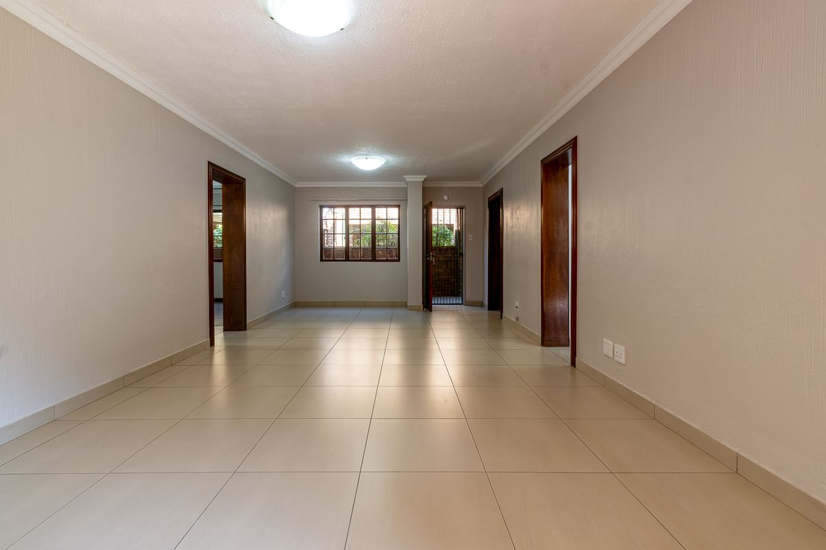 A spacious, empty living area featuring neutral tiled flooring, wooden door frames, and natural light through a rear window and security-gated door.