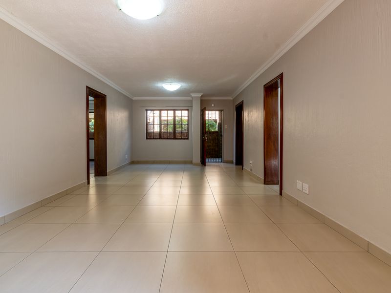 A spacious, empty living area featuring neutral tiled flooring, wooden door frames, and natural light through a rear window and security-gated door.