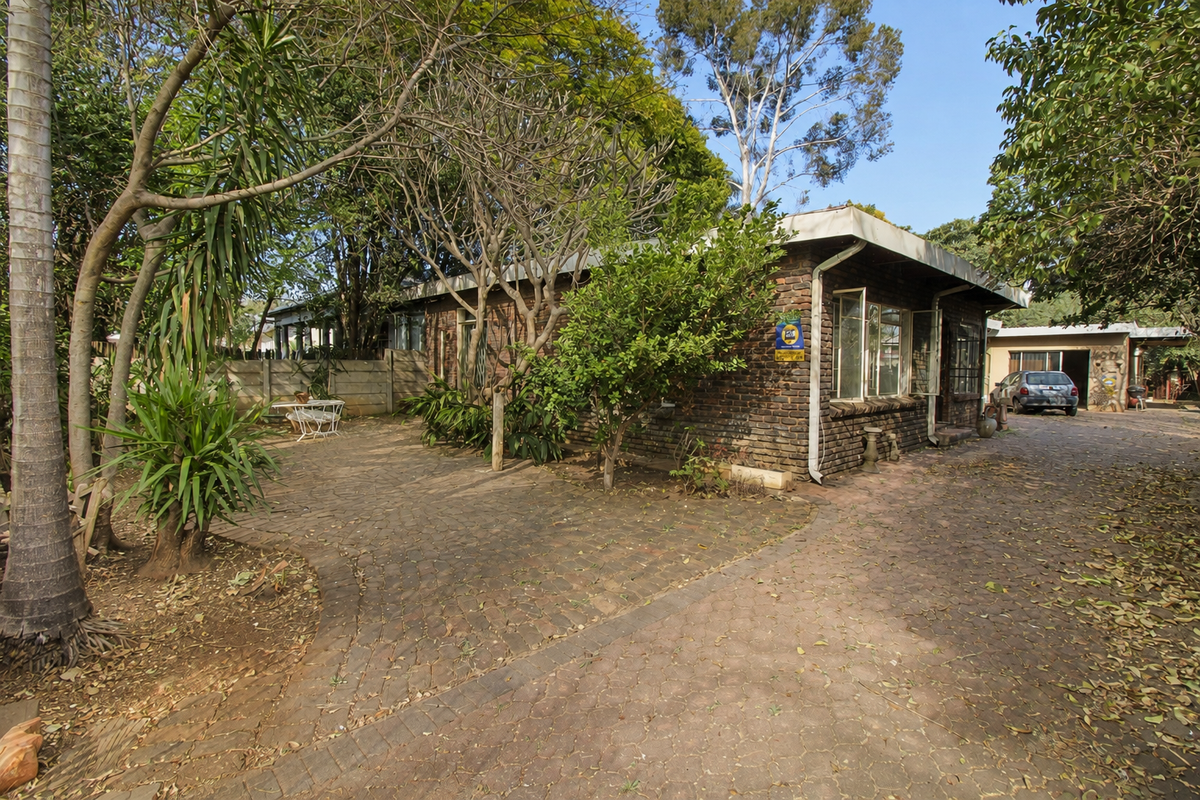 Rear view of a brick house featuring a paved driveway and patio area surrounded by mature trees and garden landscaping.