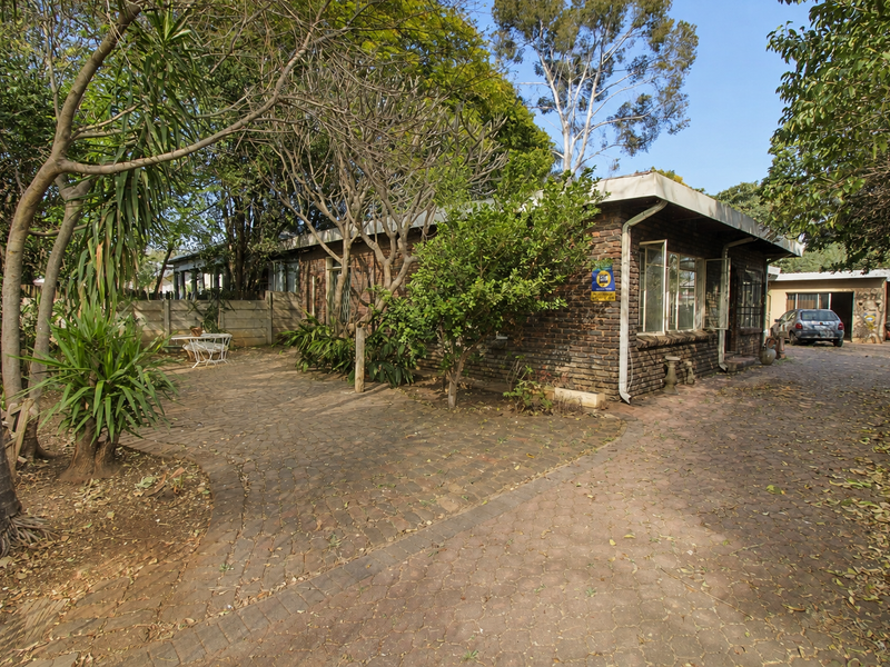 Rear view of a brick house featuring a paved driveway and patio area surrounded by mature trees and garden landscaping.