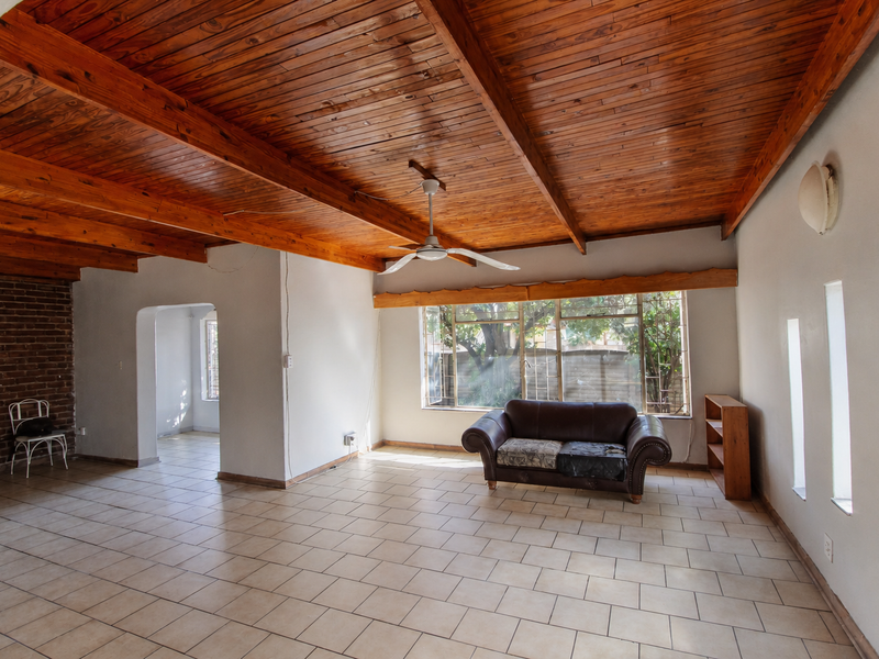 Spacious living area featuring a distinctive exposed timber beam ceiling, tiled flooring, and an exposed brick feature wall with natural light from vertical windows.