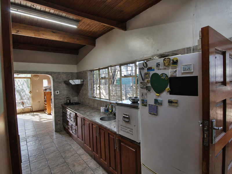 A functional kitchen featuring timber cabinetry, laminate countertops, a gas hob, and exposed timber ceiling beams, leading into an adjacent living area.