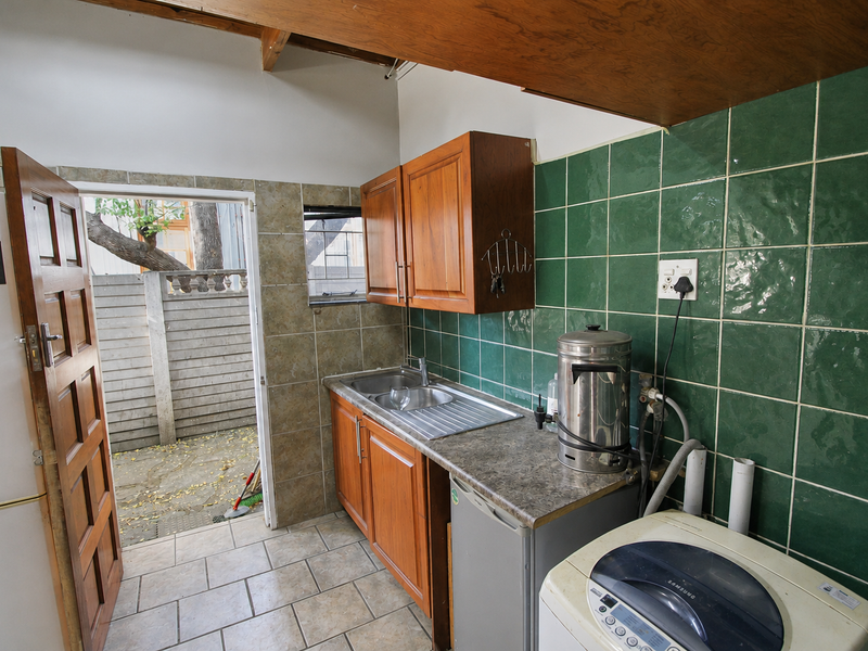 Functional laundry area featuring built-in wooden cabinetry, a stainless steel sink, green tiled splashback, and direct access to an outdoor paved area.