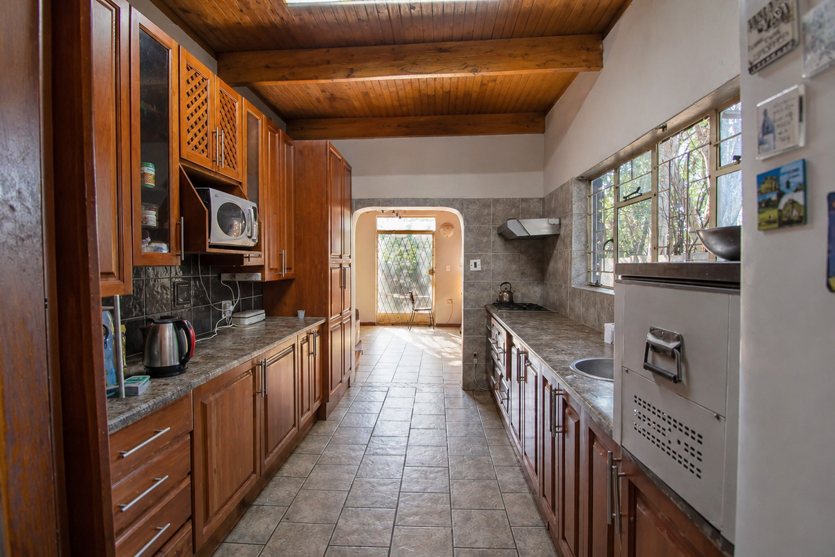 A functional kitchen featuring extensive wooden cabinetry, laminate countertops, built-in appliances, and exposed wooden ceiling beams, leading to an exterior doorway.